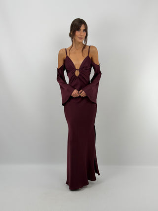 Abito lungo burgundy in tessuto satinato fluido, con top a triangolo con cut out e arricciature. Spalline sottili, spalle scoperte e maniche lunghe. Zip laterale e spacco laterale.